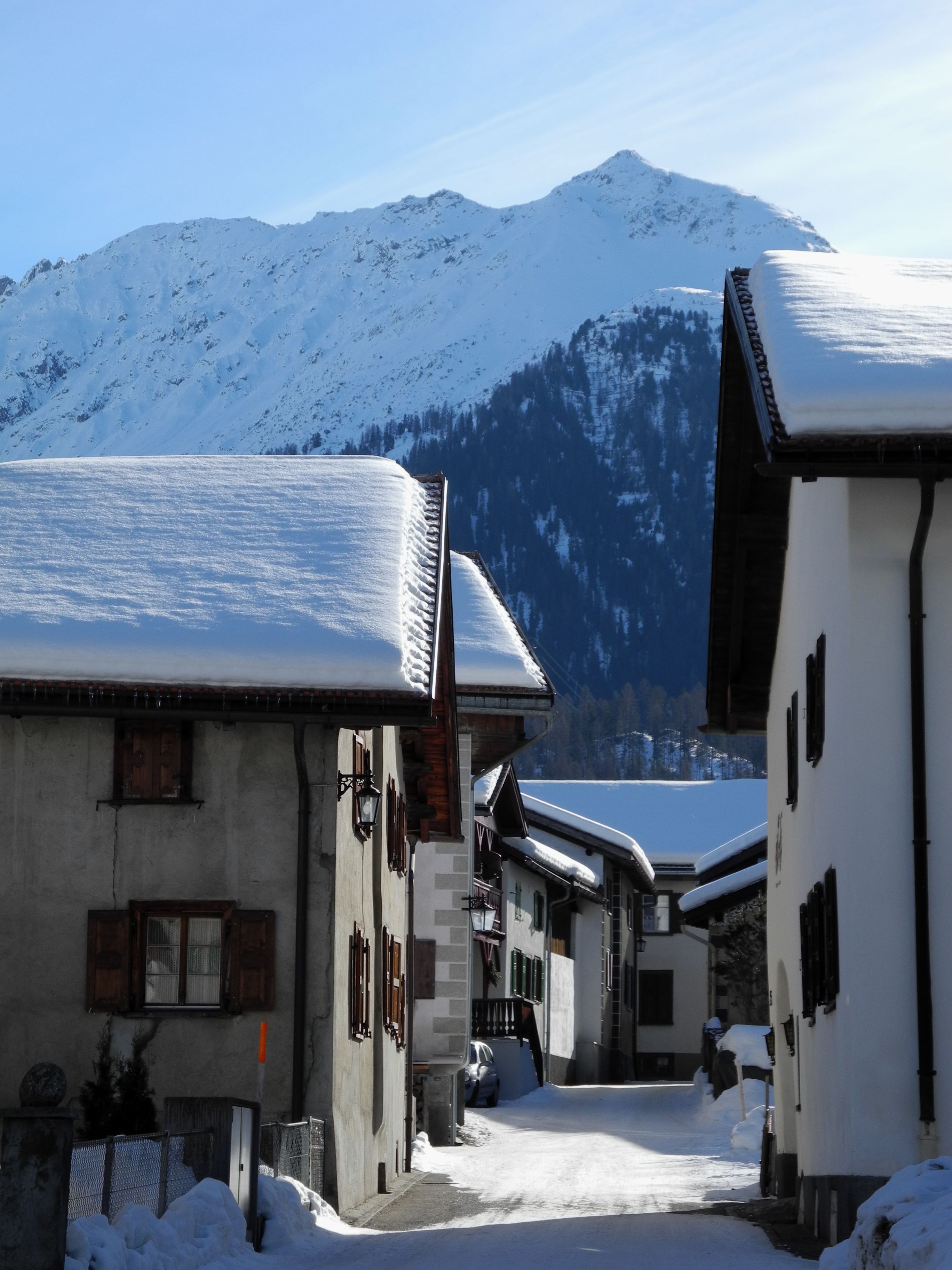 Rue enneigée de Bergün bordée de maisons engadinoises aux toits chargés de neige ; au fond, un versant montagneux enneigé sous un ciel dégagé.