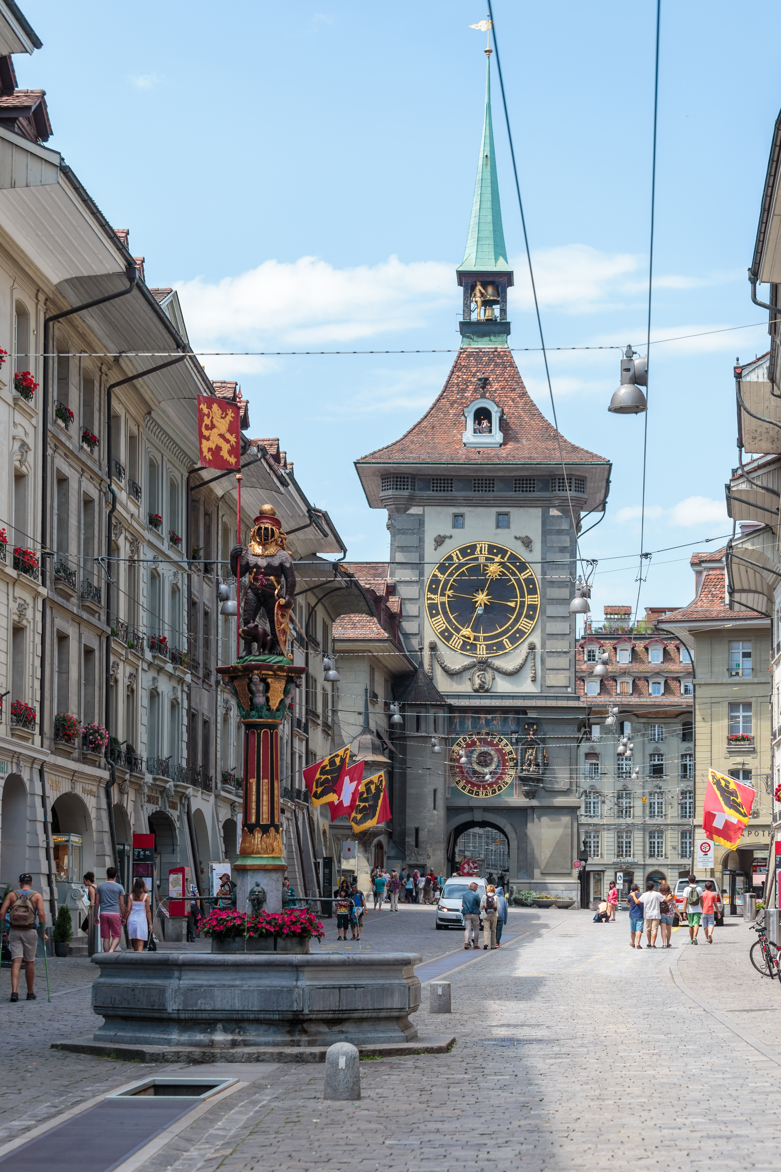 La Kramgasse à Berne, bordée d'immeubles de grès à arcades : au premier plan le Zähringerbrunnen, fontaine peinte surmontée d'un ours en armure, et au fond la tour du Zytglogge avec son cadran doré et sa flèche verte.