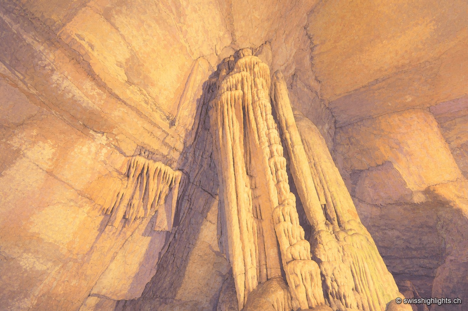 Dans une galerie souterraine éclairée de lumière artificielle chaude, un grand massif de calcite en forme de coulée figée, aux teintes ocre, vertes et mauves, s'élève du sol rocheux jusqu'à toucher la voûte de la grotte ; à sa gauche, une fine colonne stalagmitique se détache dans la pénombre, et l'obscurité naturelle de la caverne encadre la scène de tous côtés.