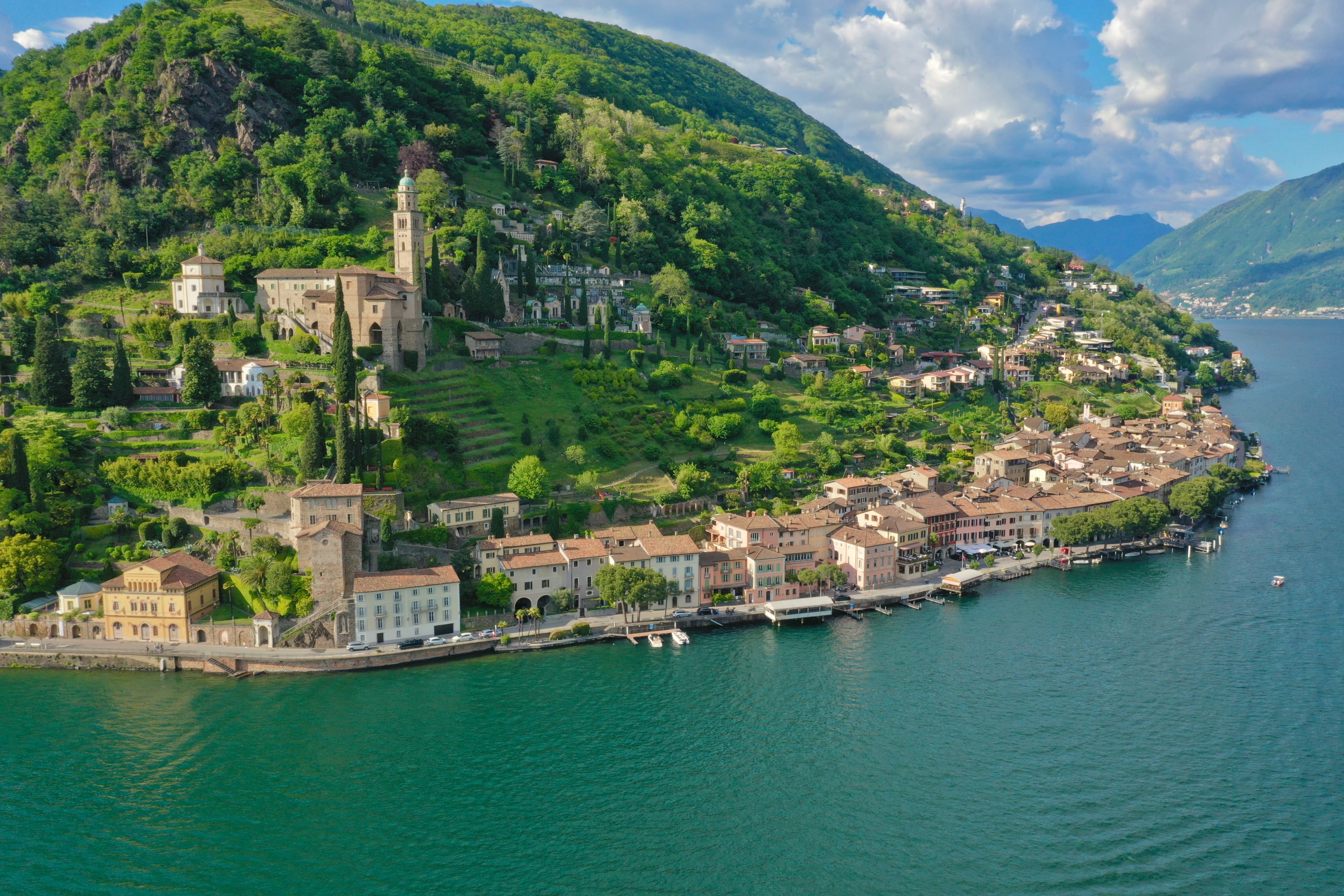 Vue aérienne de Morcote : les maisons alignées sur la rive du lac de Lugano, l'église Santa Maria del Sasso et son clocher sur le versant boisé au-dessus du village.