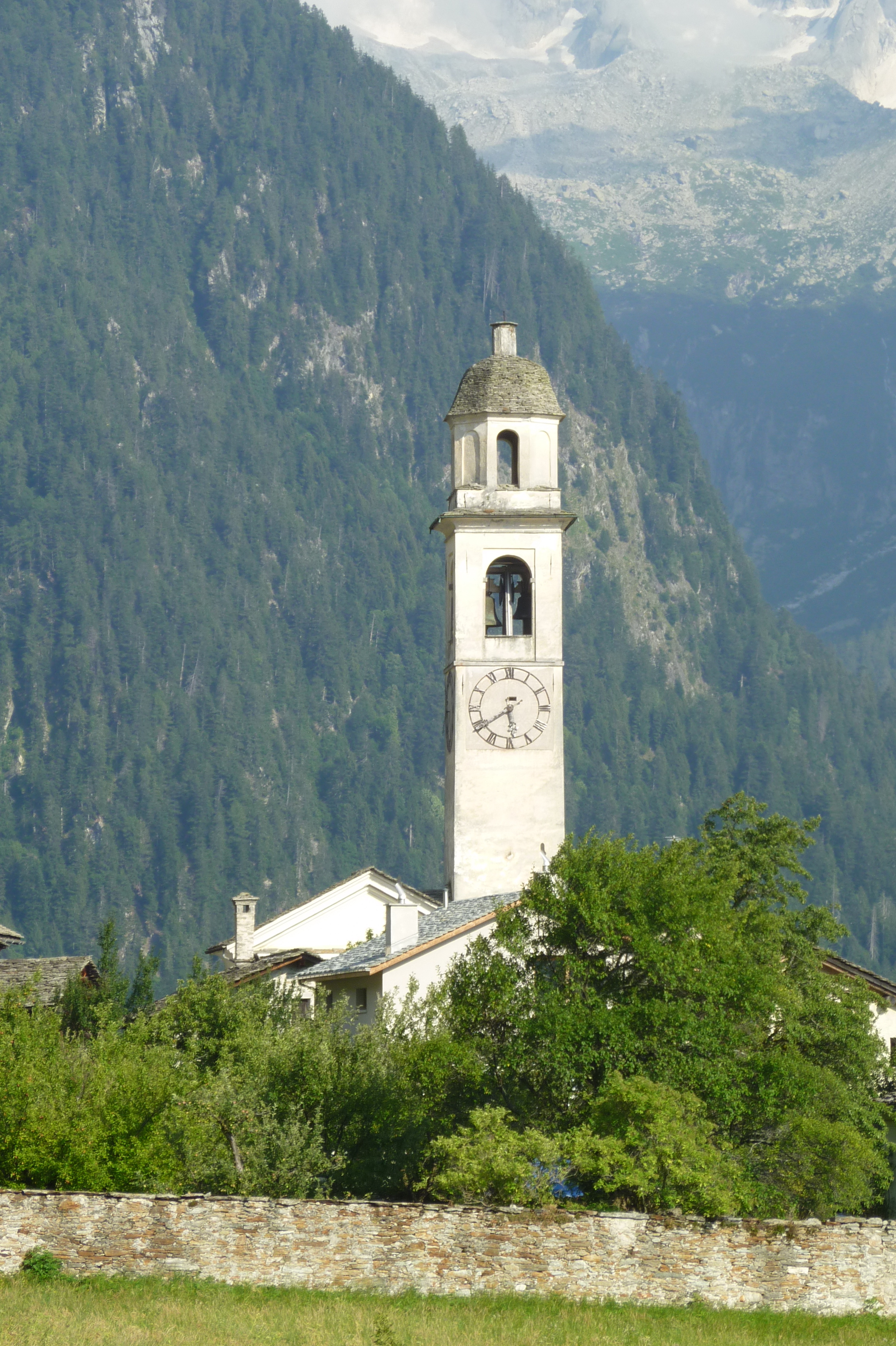 Le clocher blanc de l'église de Soglio, surmonté d'une lanterne à toiture de pierre, se détache devant un versant boisé et des parois rocheuses partiellement enneigées.