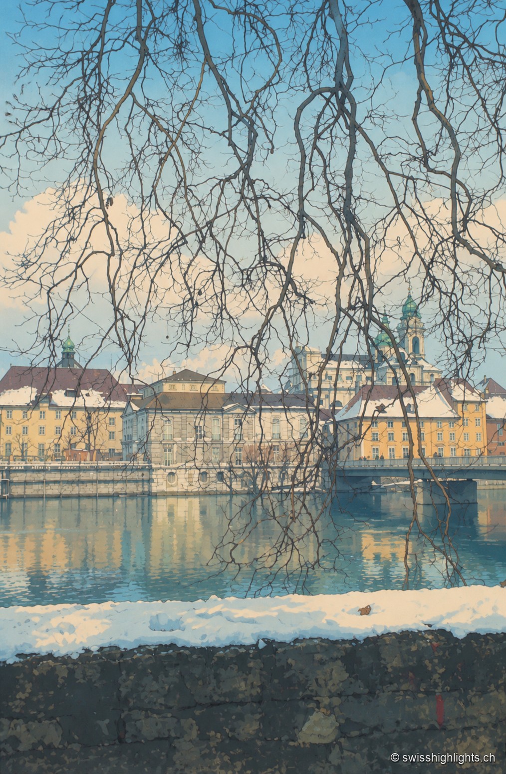 Vue hivernale de la vieille ville de Soleure depuis la rive de l'Aar : les branches nues d'un arbre encadrent les façades ocre et jaune du bourg qui se reflètent dans l'eau calme, avec un muret de pierre et une bande de neige au premier plan.