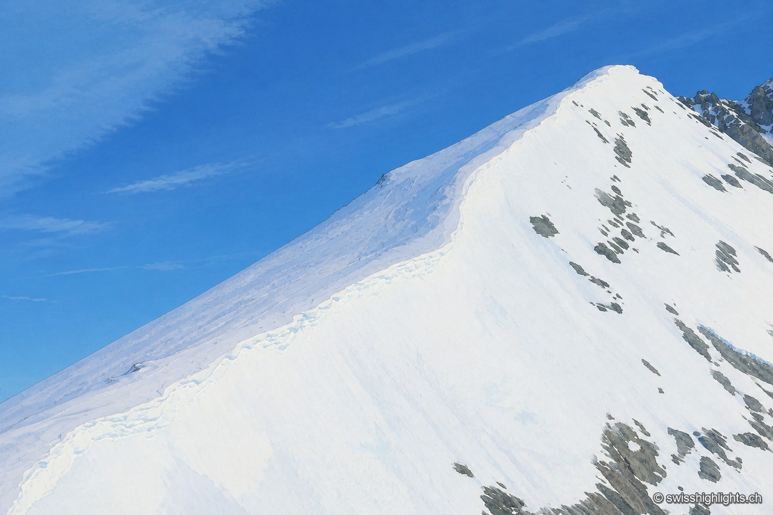 Une arête neigeuse traverse le cadre en diagonale sous un ciel bleu clair, ponctuée de quelques affleurements rocheux sombres qui percent la neige.